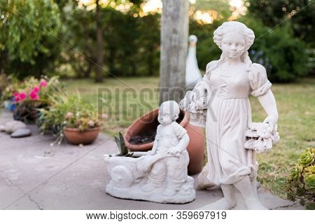 Statues In A Garden Next To Flower Pots In A Summer Day With Sunset Light