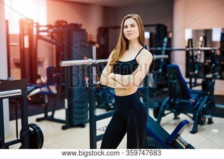 Young Beautiful Sportswoman With Nice Body. Blonde Fitness Girl Stands Cross Hands . Gym Background.