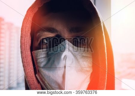 Man In A Protective Mask At Home. Sterile Medical Mask Looks Out The Window And Is At Home Quarantin