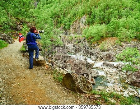 Tourist Woman With Norwegian Flag And Camera Walking Trough Mountains Path In Allmannajuvet Area Sau