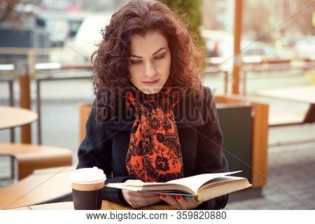A Young Blue-eyed Woman Drinks Her Drink While Reading A Book On The Terrace In Cool Sunny Weather