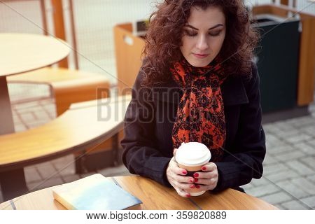 A Young Blue-eyed Woman Drinks Her Drink While Reading A Book On The Terrace In Cool Sunny Weather