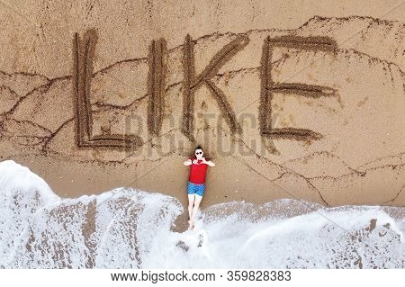 Handsome Guy In Red T-shirt And Blue Male Boxers Is Lying On Beach Washed By Foaming Sea Waves. Man 