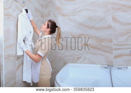 The Maid In The Bathroom Hangs A White Terry-cloth Robe On A Hanger. The Concept Of Service To Our G