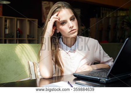 Beautiful Girl At A Table In A Cafe Is Sitting In Front Of A Laptop.