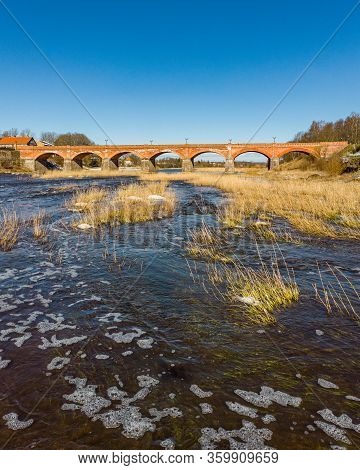 Beautiful Sunny View Of River Wenta Flowing Towards Old Brick Bridge In Old Historic Countryside Cit