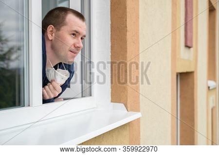 Caucasian Adult Man Smiles, Looks Out Of Apartment Window And Removes The Respirator From His Face. 