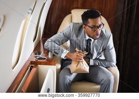 Worried African American Businessman Holding Paper Bag While Having Panic Attack During Flight On Pr