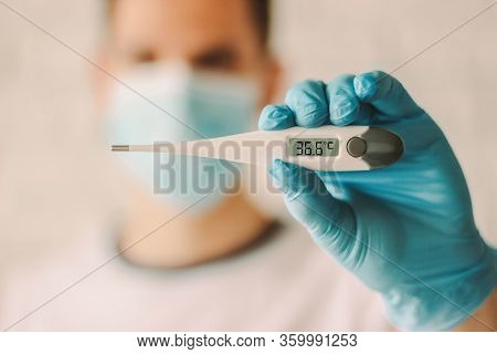 Doctor In Medical Mask And Glove Holding Electronic Thermometer In Hand. Man In Protective Mask On F