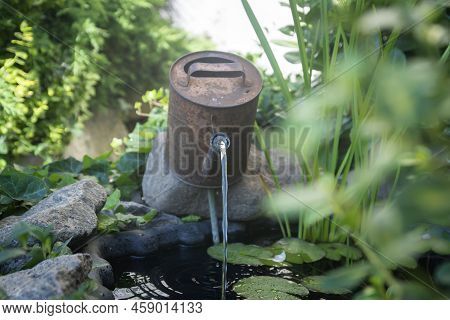 An Old, Rusty Watering Can In The Form Of A Fountain On An Artificial Pond.