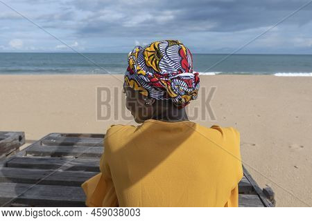 Ghanaian Woman On The Beach Wearing African Colorful Headdress And Sitting On A Wooden Bench In Keta
