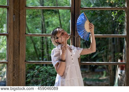Stylish Lady Wearing White Dress Hates Tropical Summer Heat. Young Blonde Woman Holds And Sways Blue