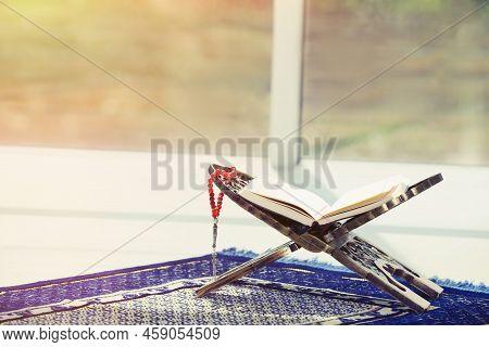 Rehal With Open Quran And Misbaha On Muslim Prayer Rug Near Window Indoors, Space For Text