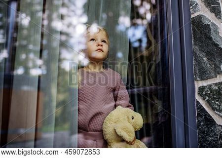 Little Child Standing Alone With Teddy Bear Behind The Window, Photo Trough Glass.