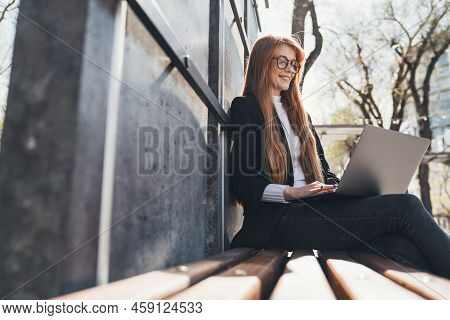 Red-headed Young Woman Sitting In Public Park Chatting Over Laptop Computer Sitting On A Wooden Benc