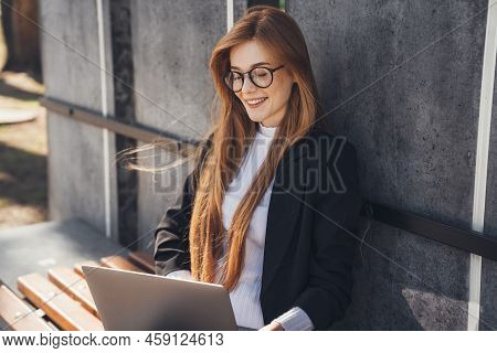Elegant Businesswoman Holding Laptop On Her Knees Sitting On Wooden Bench In Park And Working Online