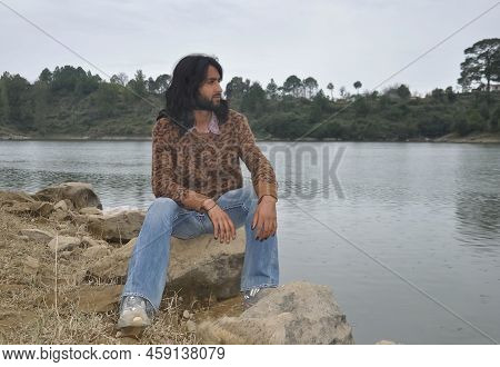 A Good Looking Young Man With Long Hair And Beard Looking Sideways While Sitting On Stone By The Lak