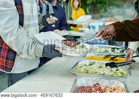 Close-up Of Volunteers Serving Homeless People And Putting Meal On Plates, They Feeding Poor People 
