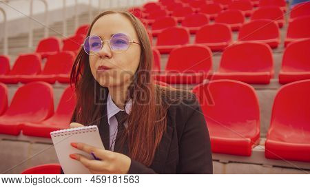 Young Woman In Glasses With Notepad, Pen Sitting On Stadium Bleachers Alone. Female Journalist Writi