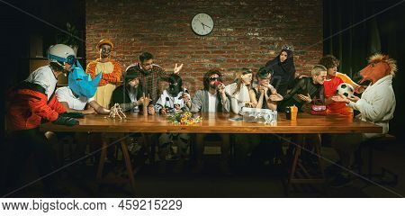 Group Of People Sitting At The Long Wooden Table In Different Costumes, Indoors