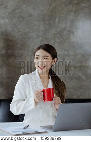 An Asian Female Employee Sits With A Red Coffee Mug In The Startup's Marketing Department Office, Sh