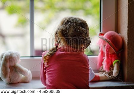Cute Toddler Girl Sitting By Window And Looking Out On Rainy Day. Dreaming Child With Doll And Toy F