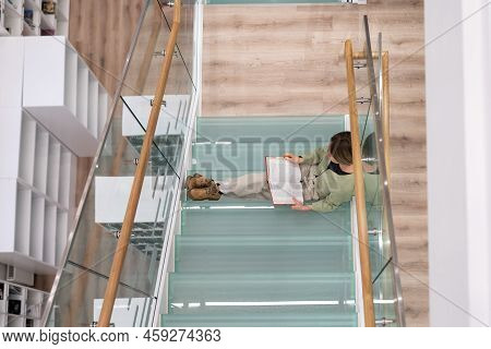 Mature Woman Sits On Glass Stairs Step In University Library Preparing For Coursework In Philosophy.
