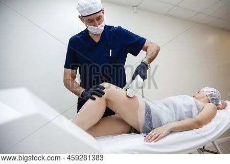 A Male Cosmetologist In A Dark Blue Uniform Conducts A Procedure Of Laser Hair Removal Of The Legs O