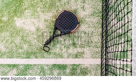 Top View Of A Black Padel Racket On A Green Court Grass Turf Behind The Net