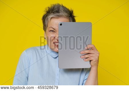 Online Troll Mature Woman Hide Behind Digital Tablet With Evil Facial Expression Isolated On Yellow 
