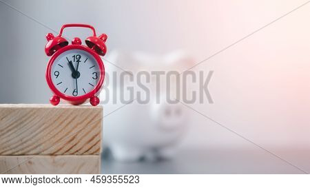 A Red Clock Rests On A Stick And Piggy Banks Both Rest On The Table. Financial And Money Deposit Con