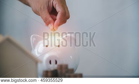 A Man's Hand Is Putting Coins Into A Piggy Bank, A Wooden House, A Pile Of Coins. All On The Table, 