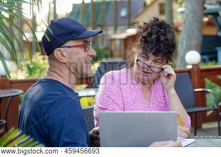 Portrait Of Middle-aged Couple. Curly Woman Looking At Smiling Man Sitting Near Laptop In Cafe Outdo