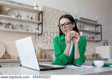 A Young Woman In Glasses And Green Clothes Is Sitting At Home In The Kitchen At The Table Watching A