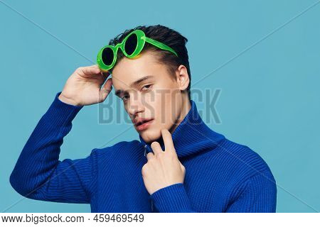 A Joyful Man Stands On A Light Blue Background In A Blue Zip-up Jacket And Holds Green Glasses On Hi