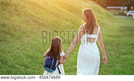 Happy Mother And Daughter First-grader Go Home After School. Woman And Girl Walk Home Jumping On Gra