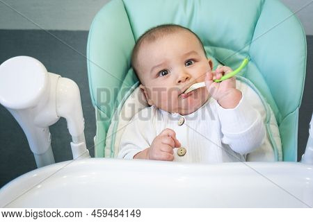 A Hungry Baby Is Gnawing On A Plastic Spoon At The Table On A High Chair. Teething, Whims, Itchy Gum