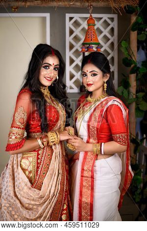 Two Indian Women Wearing Traditional Saree, Gold Jewellery And Bangles Sitting At Home With Flower A