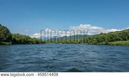 A Calm River With Lush Green Banks. Ripples On The Blue Water. Mountains On The Background Of Azure 