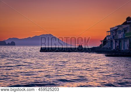 Sunset In Fishermen Village Of Klima, Milos Island, Greece. Pair Of Adults Enjoy Sunset View On Pier