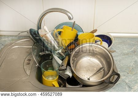 Dirty and unwashed dishes are stacked in the kitchen sink. Unwashed cups, plates, pots, forks and sp