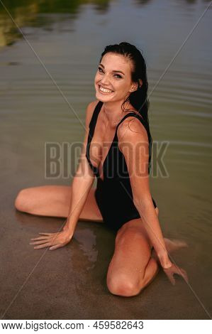 Woman In Good Mood Enjoys Life. Joyful Happy Woman In Black Piecework Swimsuit Sits On Shore In Wate