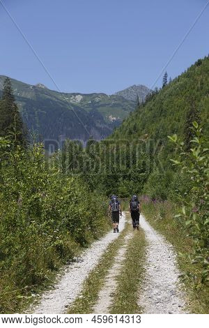 Green Valley, Mountain Peaks Against A Blue Cloudy Sky. Pedestrian Tourist - Two Man Hiking On Mount