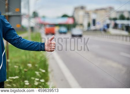 Close Up Of Mans Hand Hitchhiking By Roadside. Male Hand Showing Thumbs Up Gesture Outdoors On Blurr
