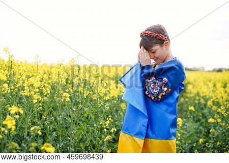 Pray For Ukraine. Child With Ukrainian Flag In Rapeseed Field. Girl Holding National Flag Praying Fo