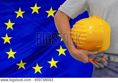 Close-up Of Hard Hat Holding By Construction Worker On Background Eu Flag. Hand Of Worker With Yello