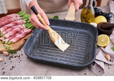 Woman Smearing Oil On A Grill Pan To Cook Aspargus Covered With Bacon