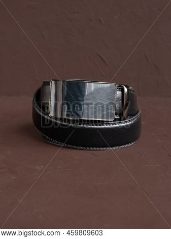 Rolled-up Black Fashionable Men's Leather Belt With Automatic Buckle On Brown Background. Male Acces