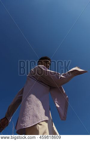 Low Anlge View Of African American Man In Striped Shirt Looking At Camera Under Blue Cloudless Sky.