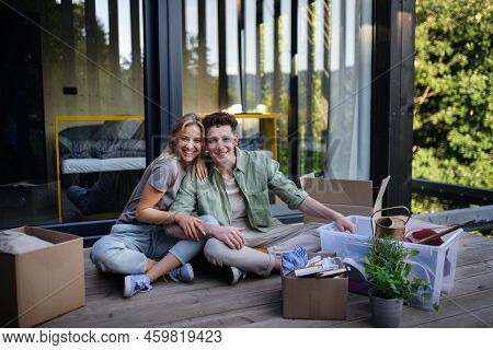 Cheerful Young Couple Moving In Their New Tiny House In Woods Sitting On Floor On Terrace. Conceptio